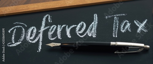 Financial term deferred tax in chalk on blackboard with pen nearby on desk
