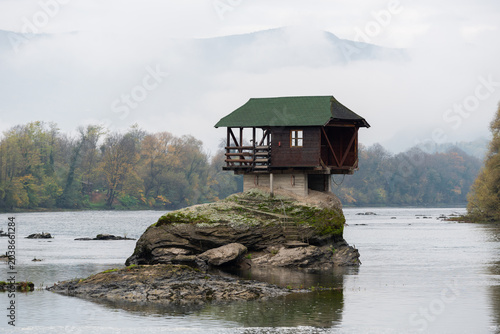 Small wooden house on rock in middle of Drina river near Bajina Basta in Serbia during cloudy morning with fog obscuring mountain in background