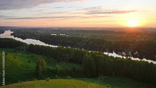 A breathtaking landscape with a winding river flowing through a dense forest and rolling green hills bathed in the warm golden light of the rising sun.
