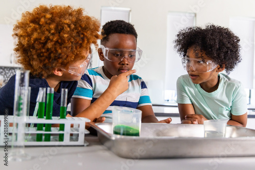Three boys sitting at school lab in short sleeves wearing safety goggles, examining test tube rack