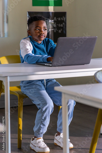 African American child working on laptop at white table in classroom, sitting on yellow chair