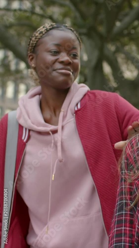 Vertical video: Approaching woman in pink hoodie smiling, hugging friend at campus with notebooks