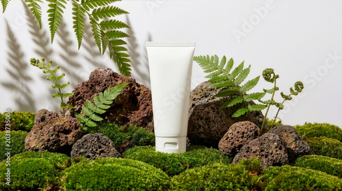 Fotografie A blank white cosmetic tube stands on lush green moss surrounded by porous rocks and fern leaves