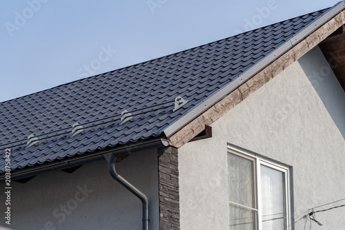 Fotografie Modern residential house exterior featuring durable dark grey metal tile roof with integrated snow guards and efficient rain gutter system against clear blue sky background