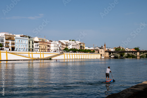 Stand up paddle boarder on Guadalquivir river with Triana bridge and colorful waterfront houses in Seville, Spain