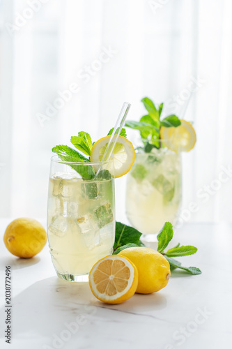 A summer drink with lemon, mint, and ice in a two glasses against a sunny window background.