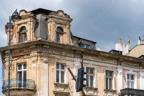 Photos Weathered facade and mansard roof of an old historic tenement house with decaying stucco, classic ornaments, windows, balcony, street lamp and traffic light under a cloudy sky