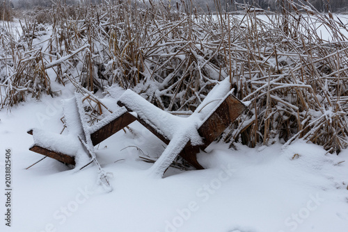 Anti-tank hedgehogs covered in snow near dry reeds next to a lake. Anti-tank defense line near the city