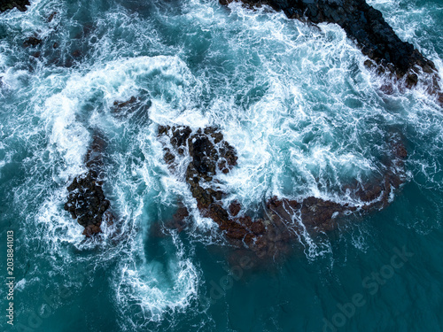 Aerial view of dark volcanic rocks surrounded by churning white sea foam and turquoise ocean waves in Caloura, Azores, Portugal.