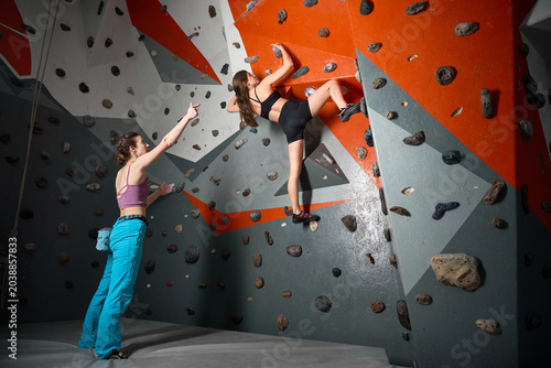 Two women share bouldering practice in modern climbing gym. Photo for support, training partnership, confidence, sport culture, wellness advertising, promotion