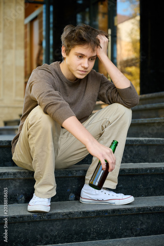 Teenage boy sits on steps with bottle in hand. Serious youth in urban setting looks withdrawn and emotionally burdened. Concept of underage alcohol risk and social concern