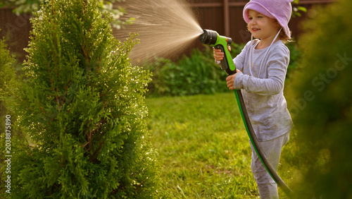 Young child wearing a hat and light clothes enjoying watering a green plant with a hose in a sunny backyard garden, helping with chores while learning about plant care