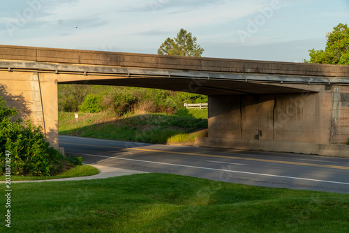 Fotografía Concrete overpass spanning a quiet road at golden hour, casting long shadows over vibrant green grass and lush trees, with a clear sky above and a small bird in flight