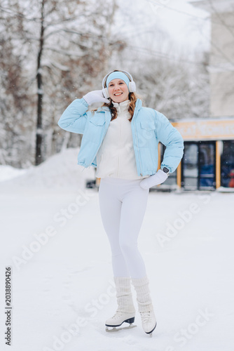 young pretty caucasian blond smiling woman in white and blue winter sport clothes skating in ice rink in winter cloudy snowy day