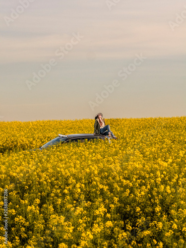 Woman Sitting on Pickup Truck Roof Surrounded by Blooming Rapeseed Field at Sunset