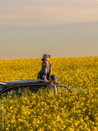 Barefoot Woman Sitting on Pickup Truck Roof Touching Face and Looking Up in Blooming Rapeseed Field