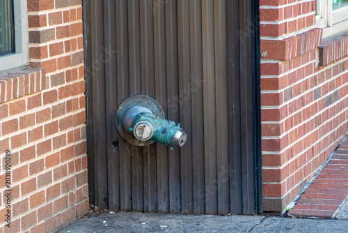 Obraz na plátně Weathered brass fire department connection standpipe with green verdigris patina mounted on a dark corrugated metal wall, flanked by an old brick building exterior and concrete sidewalk, showing