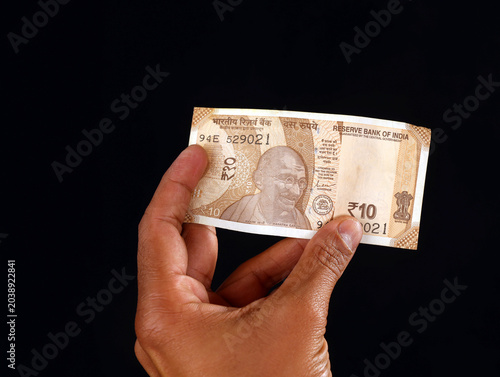 Close-up of a person holding a ten Indian rupee banknote against a solid black background, showcasing currency details and Mahatma Gandhi portrait
