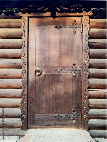 An old wooden door with metal hinges and a ring handle is installed in a traditional log house wall with carved frames. Horizontal logs and a stone base. Historic architecture and rustic style.