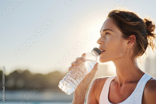 Athletic woman in her late thirties drinks water from bottle, enjoying refreshing moment outdoors. sunlight highlights her features, conveying sense of vitality and health