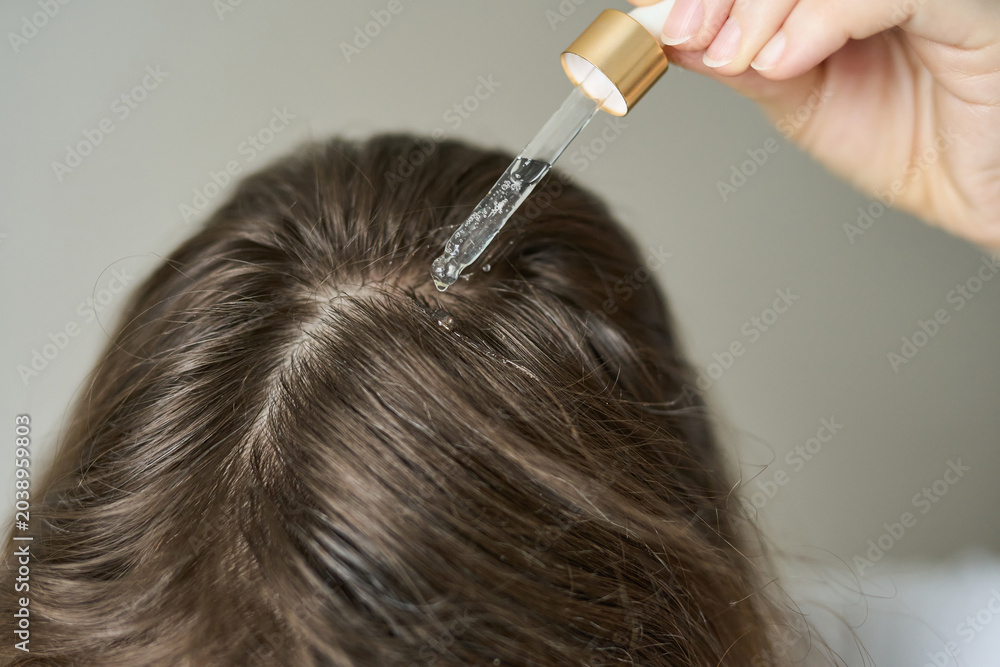 Fototapeta premium Woman Placing Clear Liquid Drops From Glass Pipette Onto Scalp Along Center Part Of Brown Hair In Closeup Indoor Grooming Scene Showing Precise Topical Application And Parted Roots.