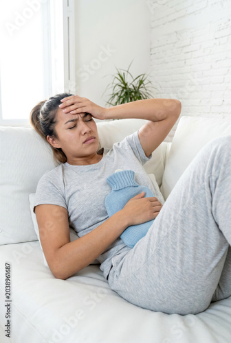 Young woman wearing gray pajamas lying on white sofa with blue hot water bottle, suffering from menstrual cramps.