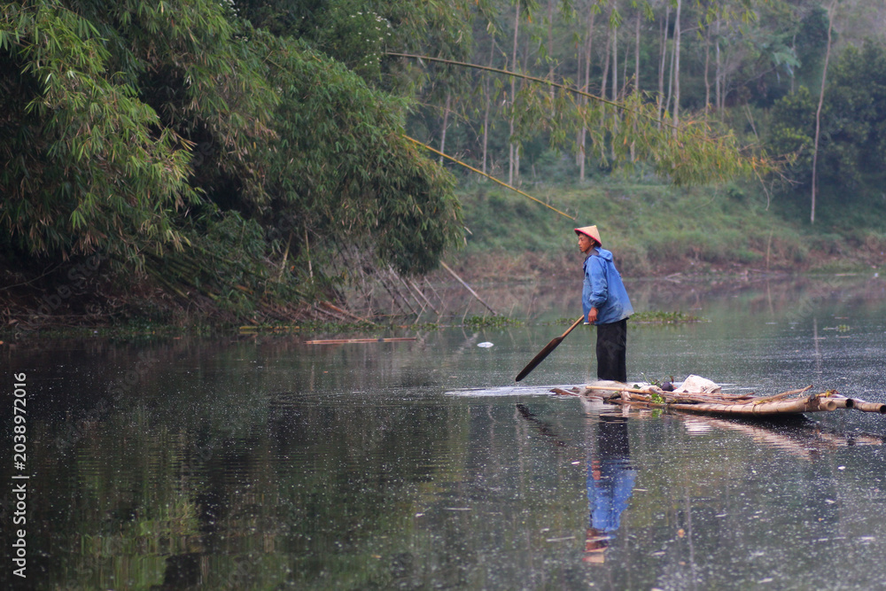 Naklejka premium Asian Indonesian male fisherman navigating a traditional bamboo raft on a calm river surrounded by lush nature.
