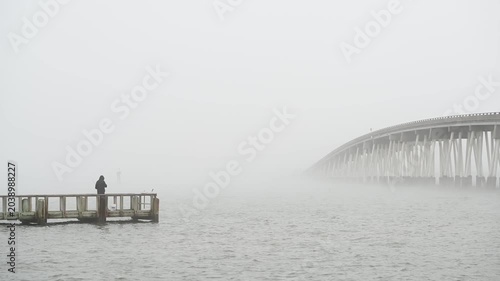 A distant fisherman stands on a pier casting a line into calm waters at Assateague Island, while a bridge on the right fades into thick fog, creating a quiet and atmospheric coastal scene.