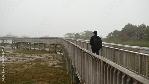 A photographer carrying a camera backpack walks along a wooden boardwalk through fog-covered wetlands at Assateague Island, Maryland, surrounded by a quiet and atmospheric coastal landscape