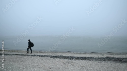 A lone photographer carrying a camera and tripod walks along a misty, deserted shoreline, capturing the quiet atmosphere and sense of exploration on a fog-covered beach.