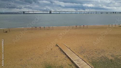 Drone footage rising from the sandy shoreline of Sandy Point State Park in Maryland, gradually revealing the Chesapeake Bay Bridge stretching across the distant waters of the Chesapeake Bay
