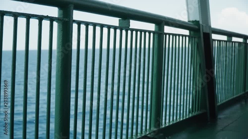 Ferry railing overlooking calm blue ocean, rusted green metal balustrade framing steady horizon, gentle waves lapping, soft daylight, cinematic minimal composition, deck planks and safety post,