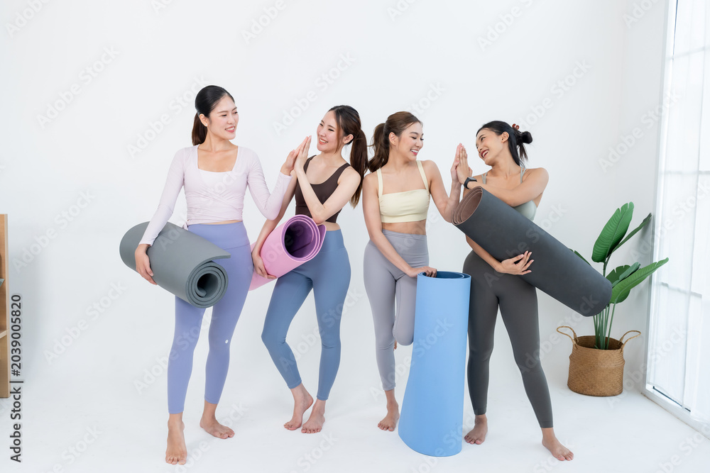 Naklejka premium Asian women friends hold colorful yoga mats and share high five in bright studio, fitness community celebrates teamwork, wellness motivation, flexibility progress, posture balance, healthy habit