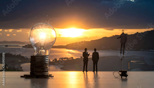 Business team observing a glowing lightbulb on stacked coins, symbolizing innovation and financial growth during a scenic sunset overlooking the ocean