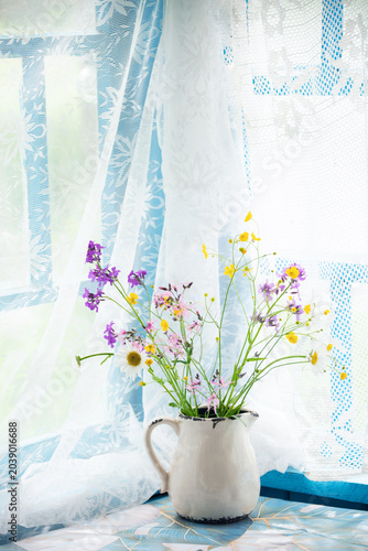 Wildflowers in a rustic pitcher on a windowsill with lace curtains. A charming arrangement of delicate wildflowers in a vintage ceramic pitcher sits on a windowsill, bathed in soft natural light