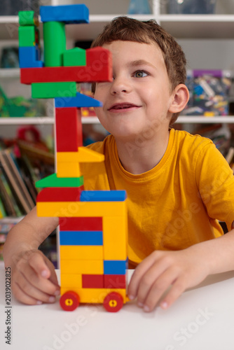 Laughing preschool boy in yellow shirt sitting beside tall colorful toy block tower on wheels at white table in bright children's playroom