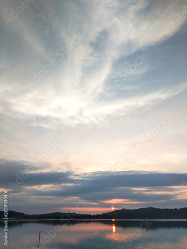 The view of the sun rising from behind the hill reflected on the surface of the lake with a beautiful sky of thin clouds.