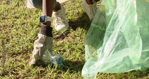 African American woman spotting bottle on grass squatting placing bottle into green bag for cleanup