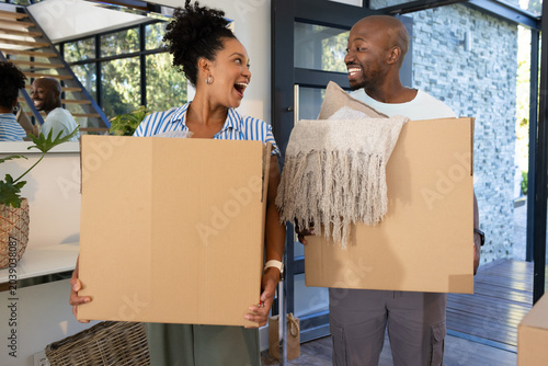 African American couple carrying cardboard moving boxes with blanket, pillow smiling in home foyer
