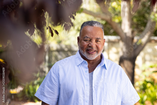 Senior African American man standing in sunlit backyard wearing light blue shirt, framed by leaves