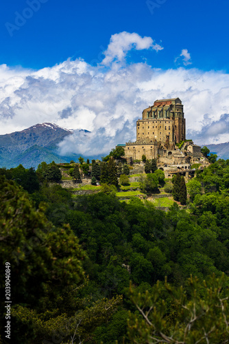 Sacra di San Michele abbey perched on Mount Pirchiriano at the entrance of the Susa Valley in spring, with the still snow-capped Alpine peaks emerging through the clouds in the background