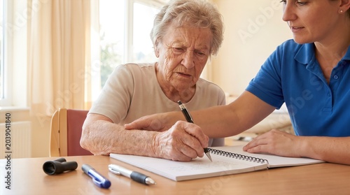 Senior woman writing in notebook with caregiver assistance. Elderly lady using pen grip aid. Cognitive training and handwriting support for seniors