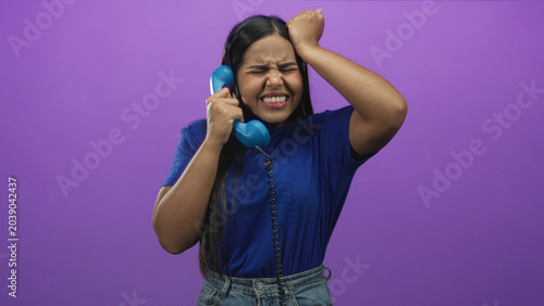 Young hispanic woman in blue tshirt holds blue handset telephone, gestures angrily with raised hand and clenched face in studio; frustration tension.