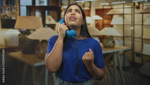 Foto Woman holding blue handset to ear, eyes closed with fist raised and open hand gesture in a lamp showroom building; melancholy longing