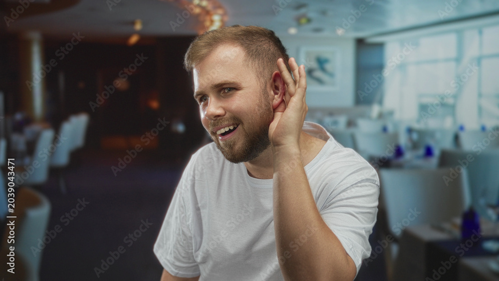 Obraz premium Man cupping hand to ear listening in building dining area wearing white t shirt and beard; curiosity listening engagement.