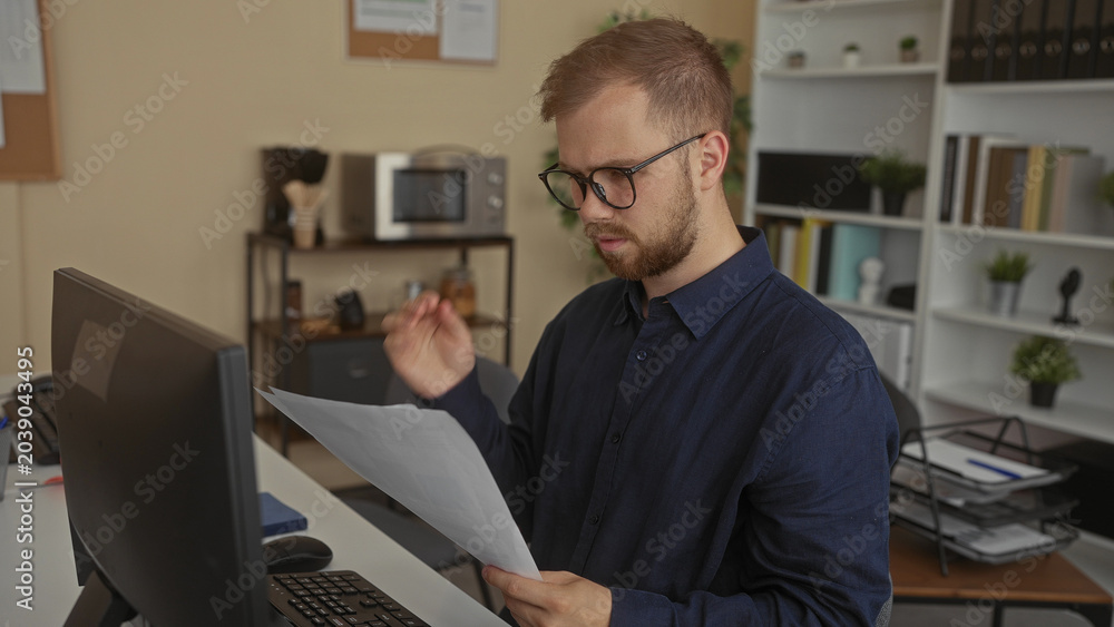Fototapeta premium Man reading printed document and holding paper at a building office desk near computer monitor and shelves; concentration paperwork.