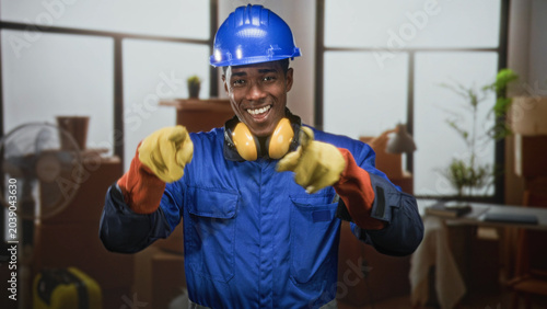 Φωτογραφία Man in blue hardhat and coveralls smiling with yellow ear defenders and heavy gloves points forward with gloved hands in building; confidence safety teamwork