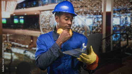 Quadro em tela Man in hardhat wearing blue coverall and noise cancel headphones holding laptop with wrench and hand to chin in building; focused problem solving