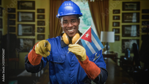 Photography Man in hardhat holding cuban flag, wearing yellow gloves and earmuffs, smiling while pointing finger toward camera in building; national pride