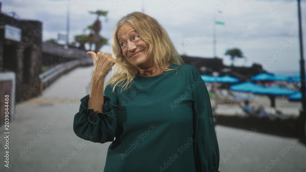Obraz premium Senior woman points thumb to street beside beach umbrellas while smiling and showing hand; contentment.